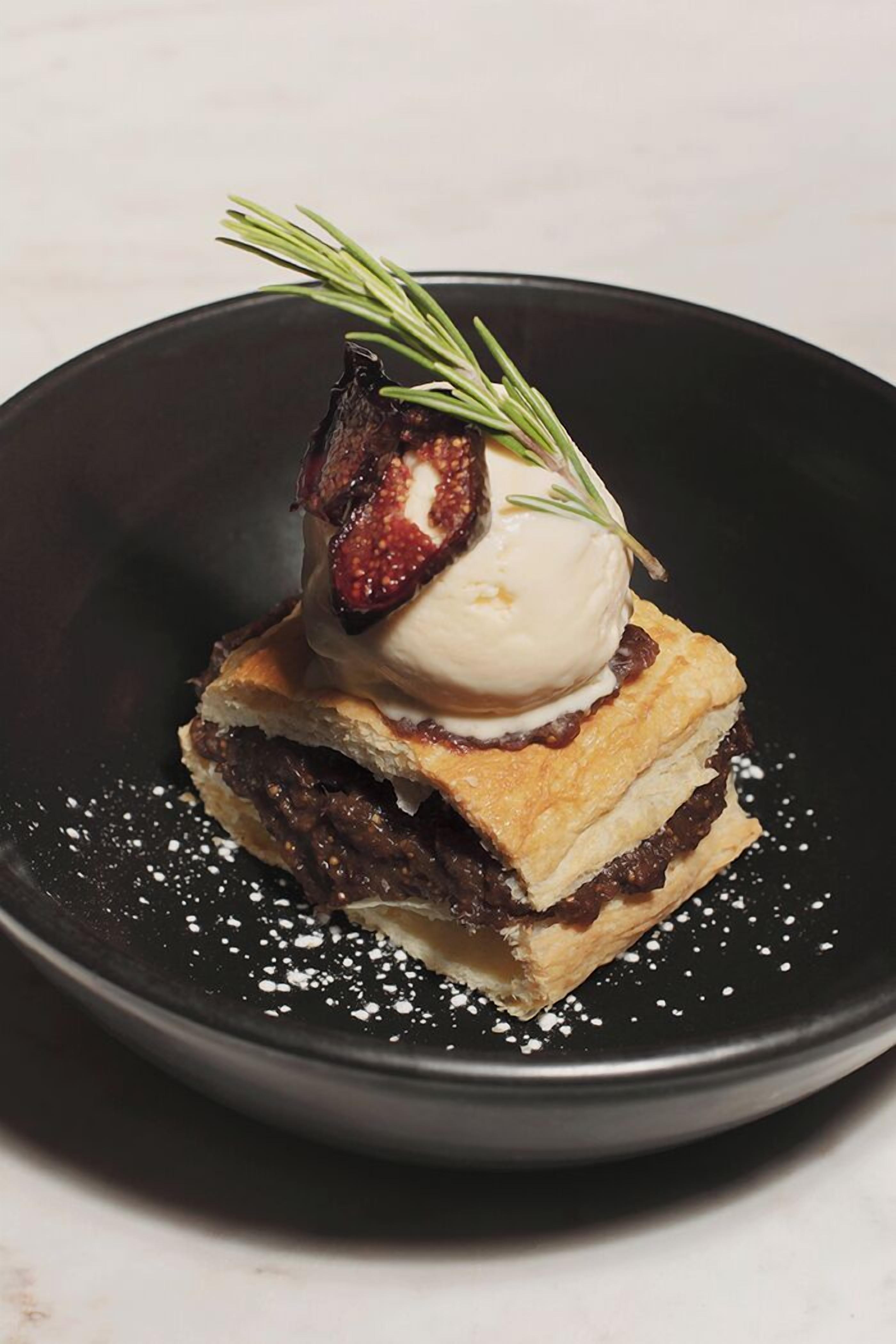 Un postre con fruta, helado y una ramita de romero sobre un plato negro.