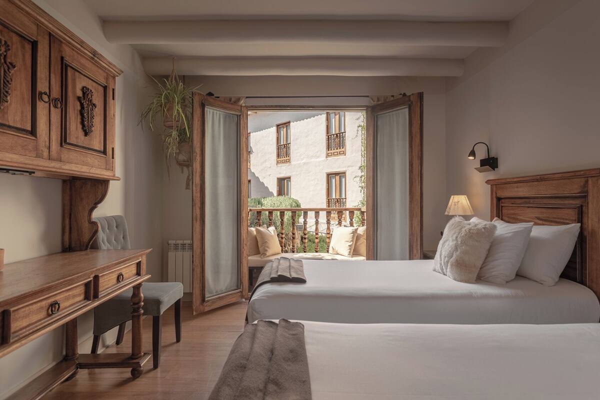 A hotel room with two beds, a desk, a chair, a window with a view of a building, and a balcony.