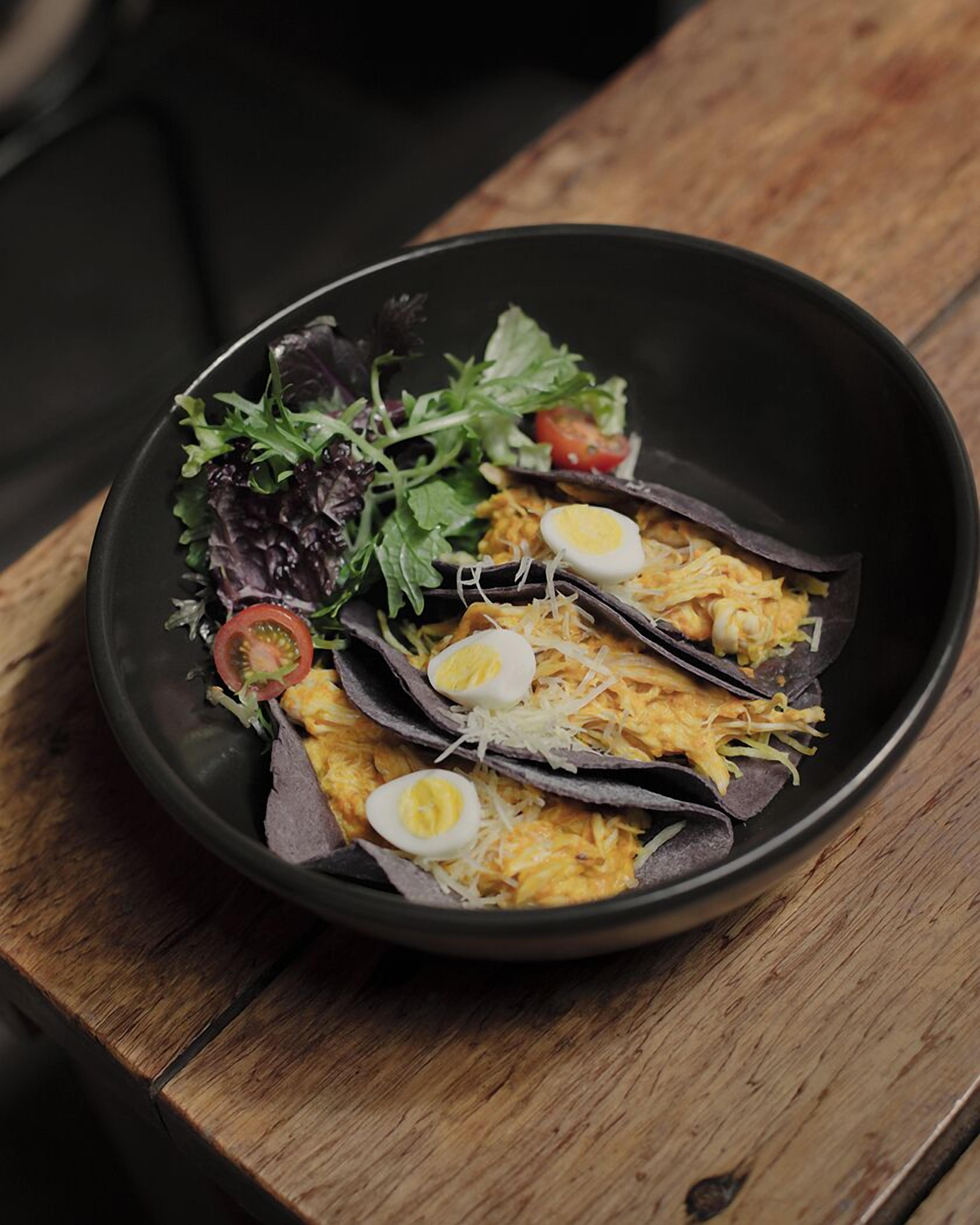 A black bowl containing a meal with tortillas, eggs, shredded cheese, and a side salad.