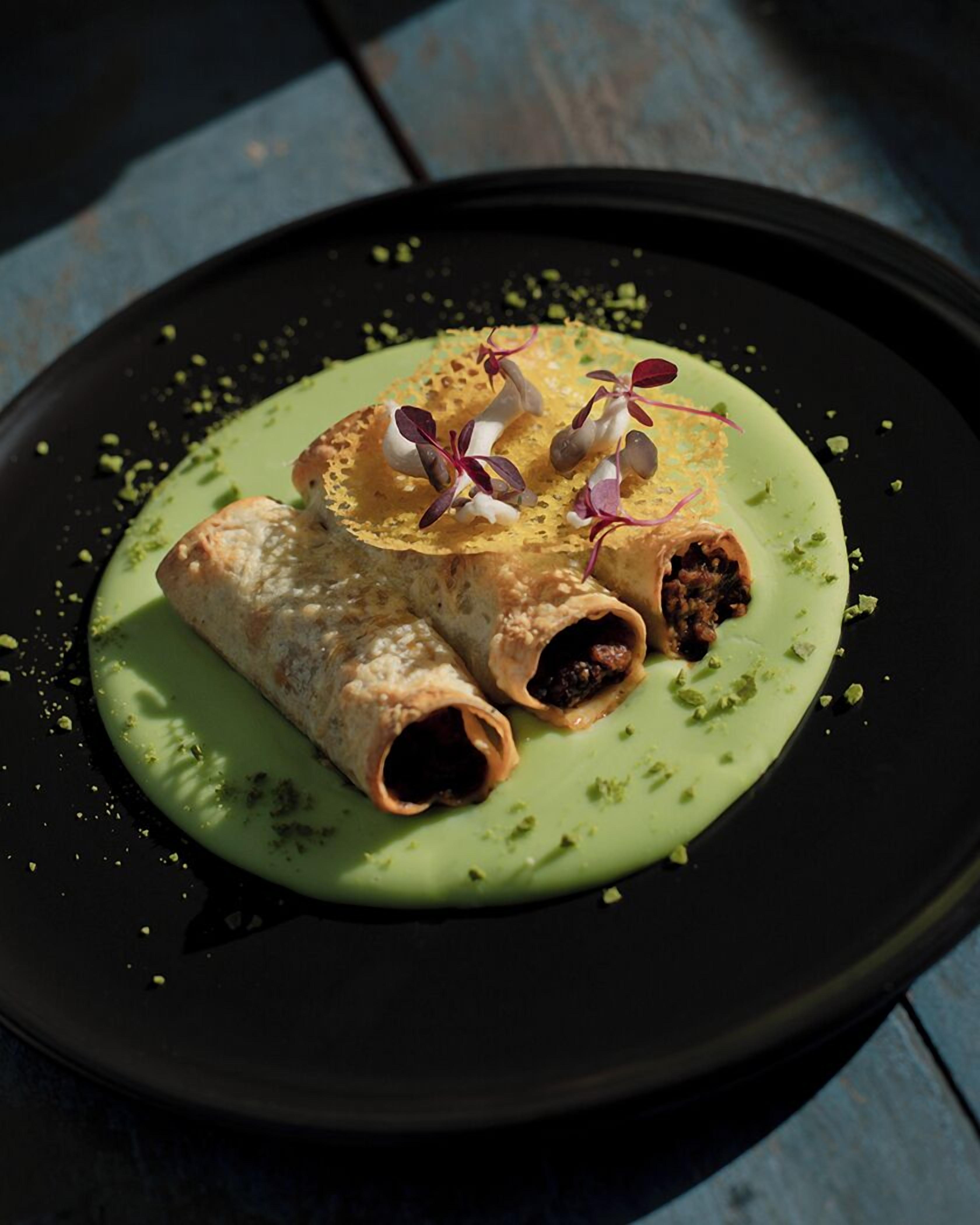 A plate with rolled items, possibly food, on a dark plate with green sauce and garnishes.