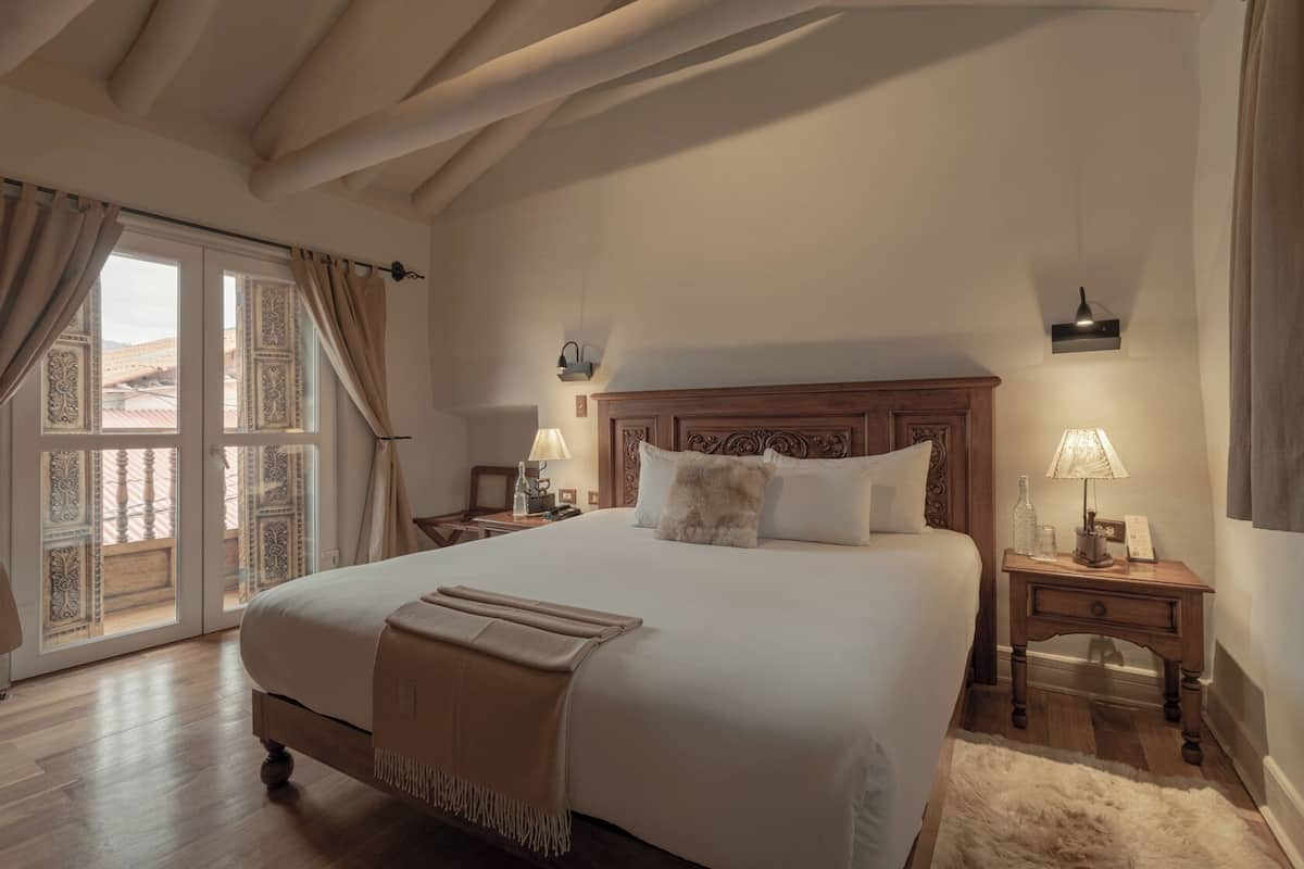 A spacious bedroom with a large bed, wooden headboard, bedside tables with lamps, and a view through a glass door.