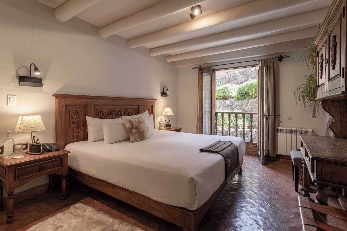 A spacious bedroom with a large bed, wooden headboard, bedside lamps, a nightstand, and a view of a stone wall and greenery outside.