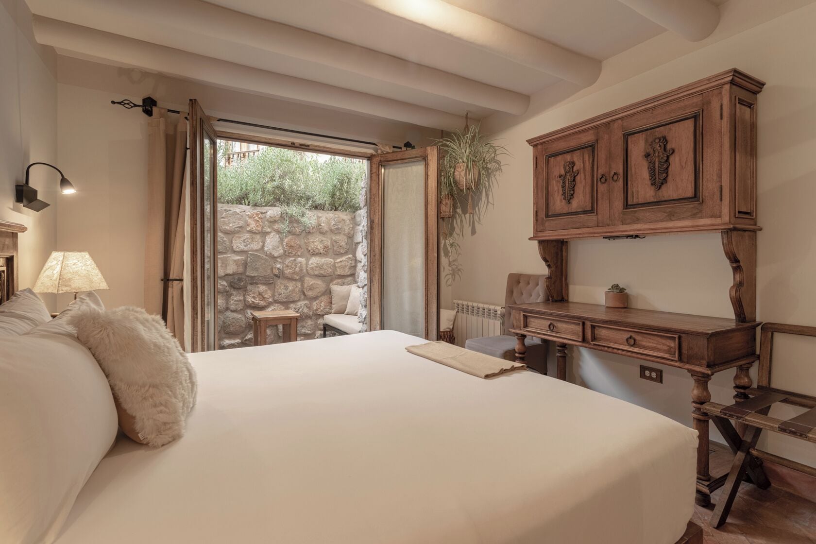 Un dormitorio con una cama grande, un aparador de madera, un escritorio, una lámpara y vistas a una pared de piedra y follaje exterior.