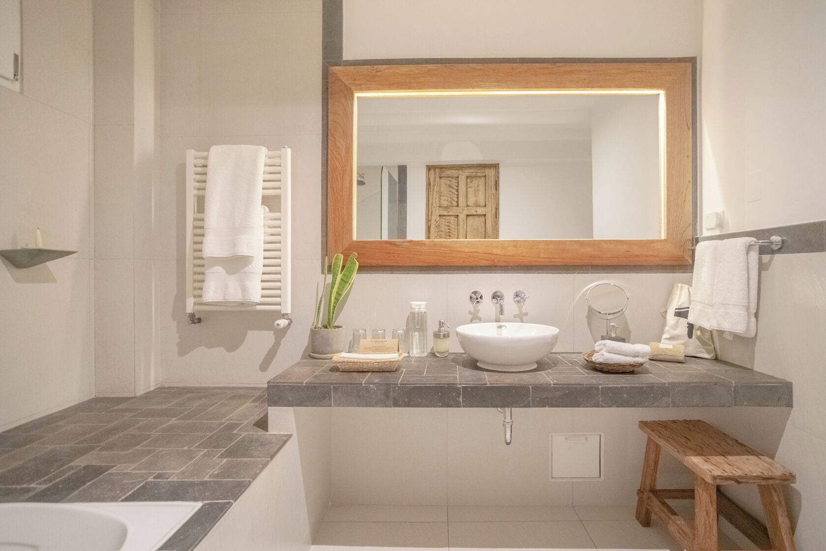 A bathroom with a white sink, a wooden-framed mirror, a towel rack, a small wooden stool, and a potted plant.