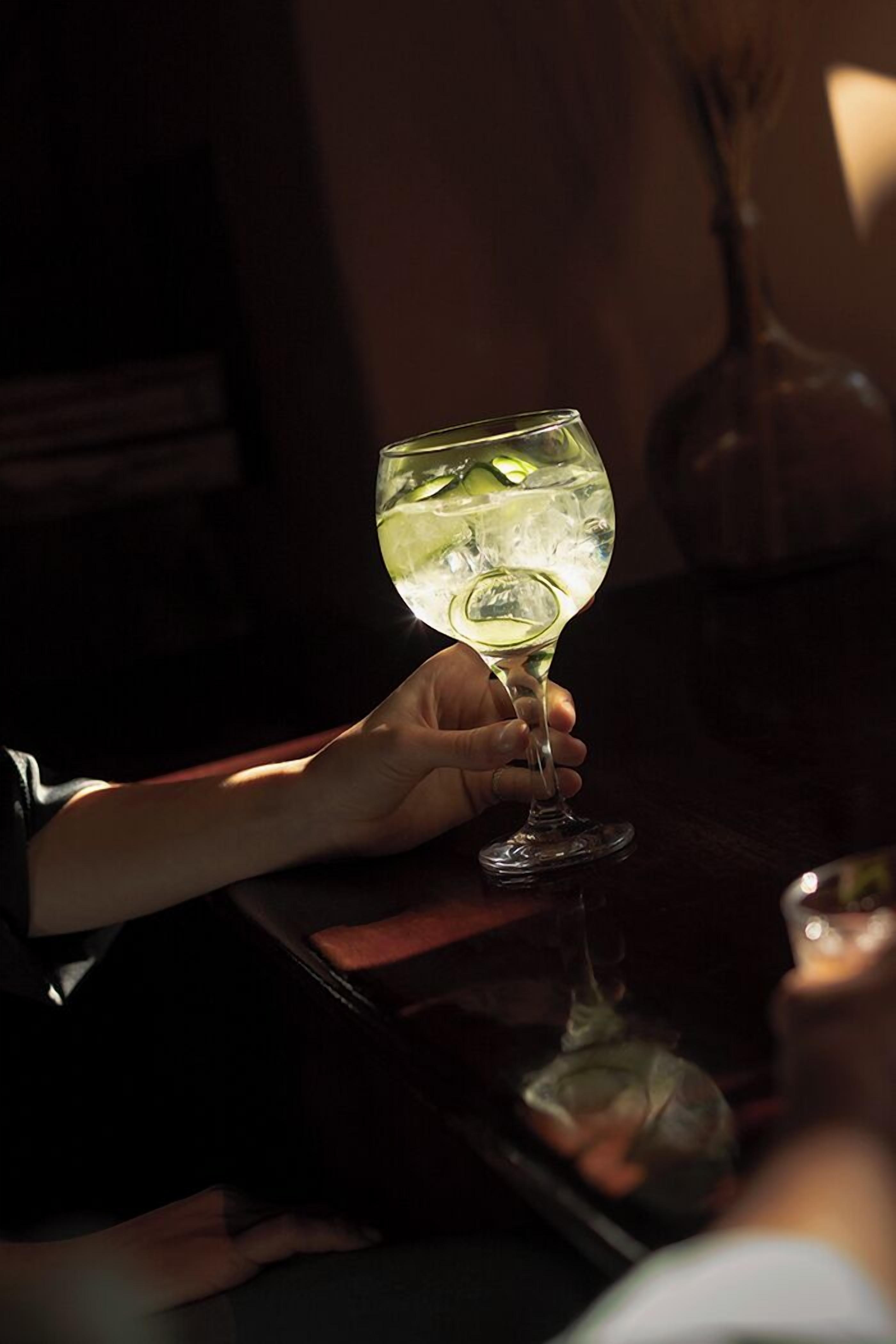 A person holding a wine glass with ice cubes, seated at a table with a dark wooden surface.