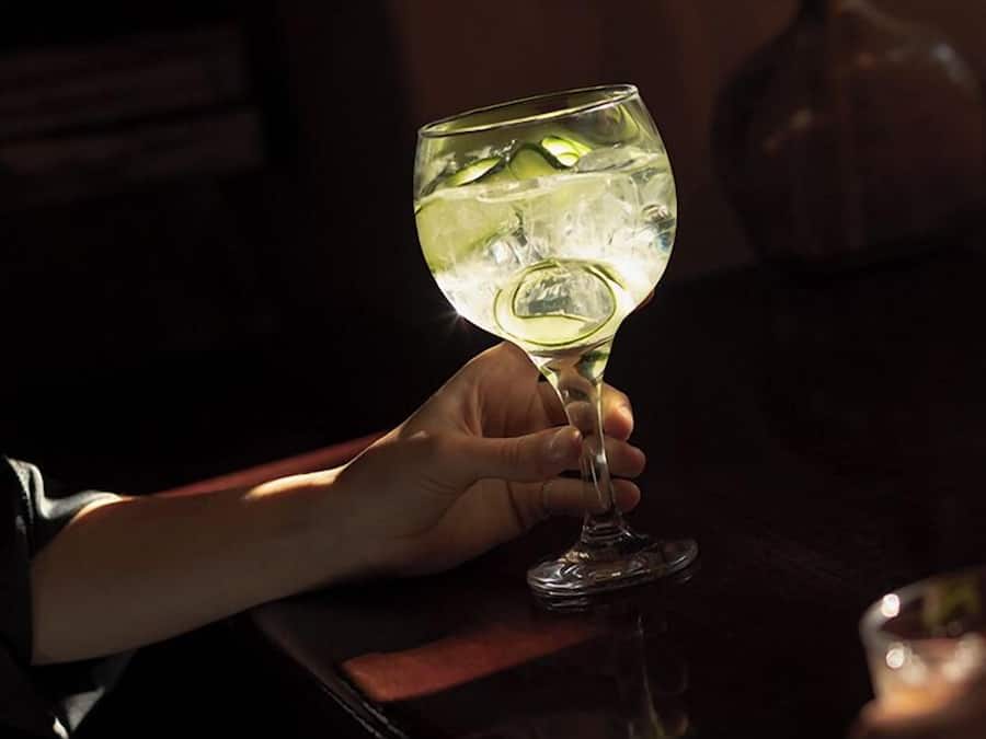 Una persona sosteniendo una copa de vino con cubitos de hielo, sentada a una mesa con superficie de madera oscura.