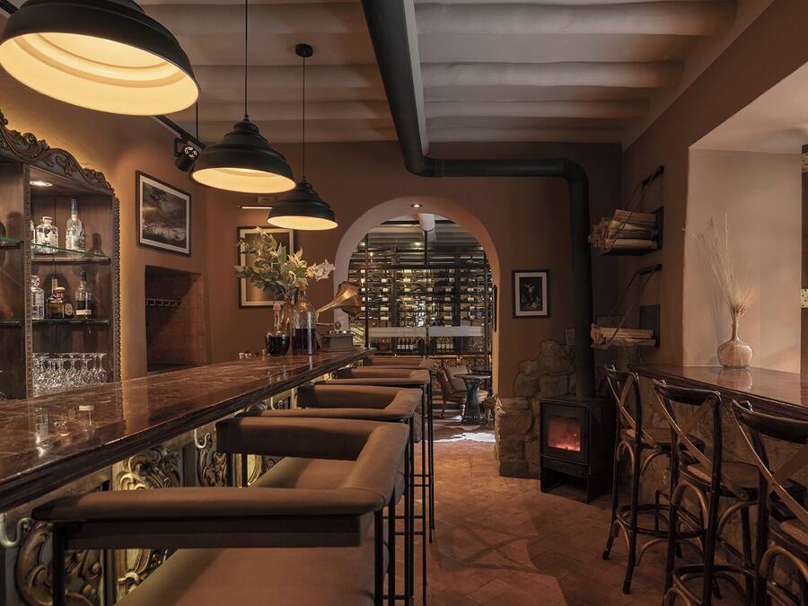 A bar area with a long counter, multiple bar stools, and hanging pendant lights. There is a stone fireplace and a bookshelf filled with books and decorative items.