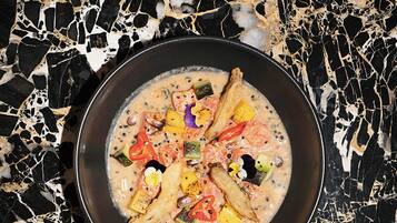 Un tazón de sopa con diversos vegetales y carne sobre un plato negro, colocado sobre una superficie de mármol blanco y negro.