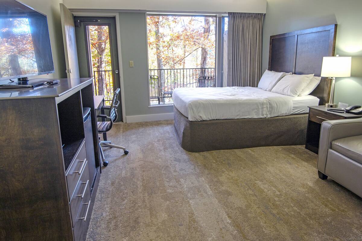 A hotel room with a bed, a desk, a chair, a sofa, a TV, a balcony with a view of trees, and a window with curtains.
