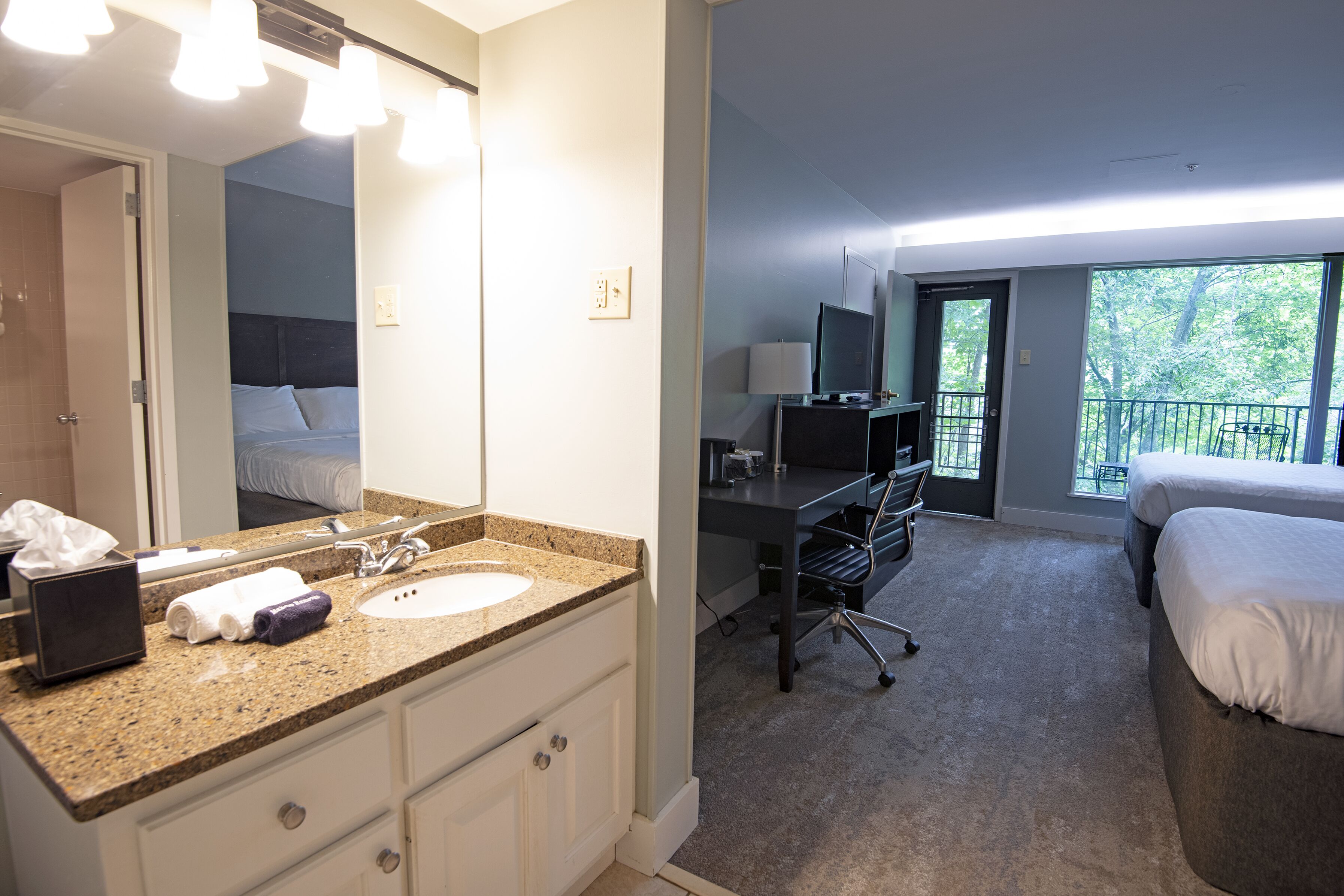 A hotel room with a large bathroom vanity, a desk with a chair, a bed, and a balcony with a view of trees.