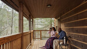 A wooden deck with a railing, a person sitting on a chair, and another person standing and looking through binoculars. - Natural Bridge State Resort Park (Slade)