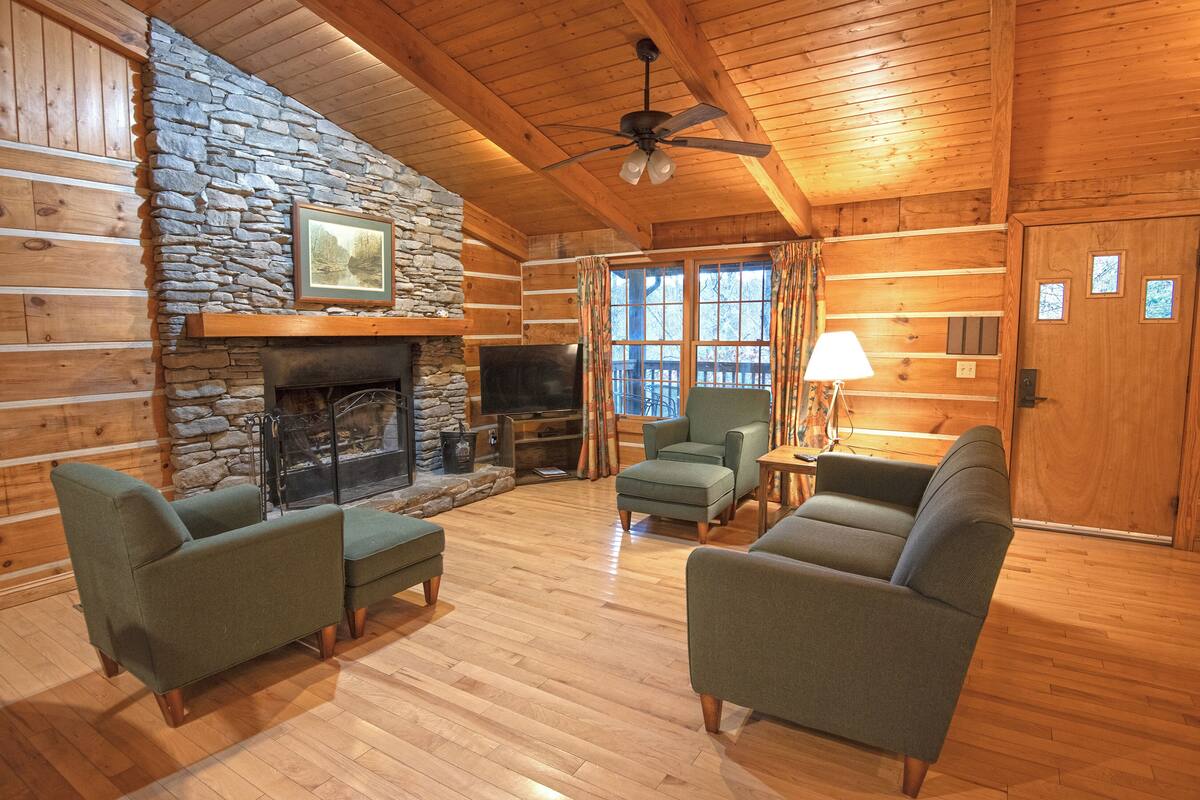 A log cabin interior with a stone fireplace, wooden walls, and a ceiling fan.