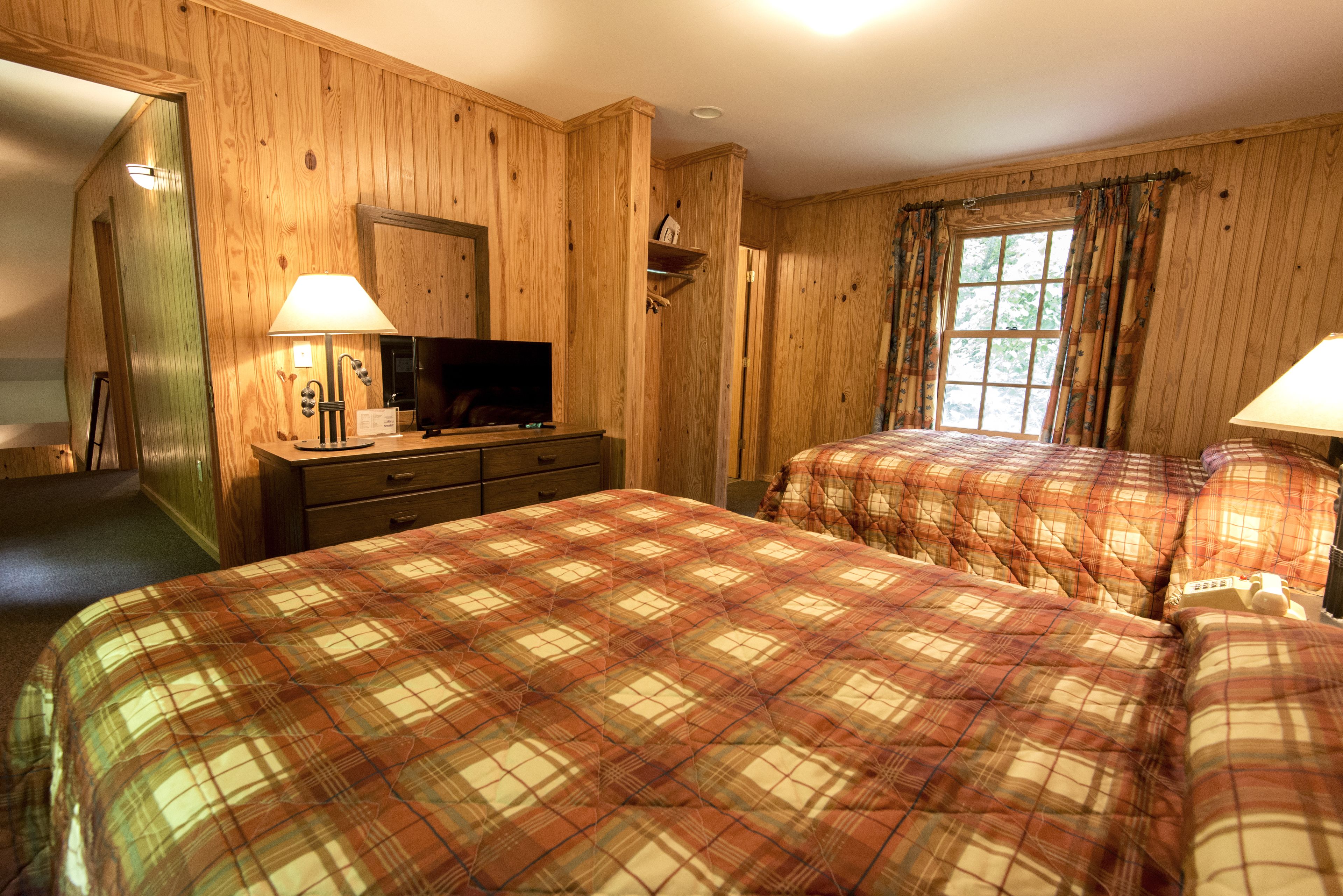 A room with wooden paneling, two beds with plaid bedding, a television on a dresser, and a window with curtains.