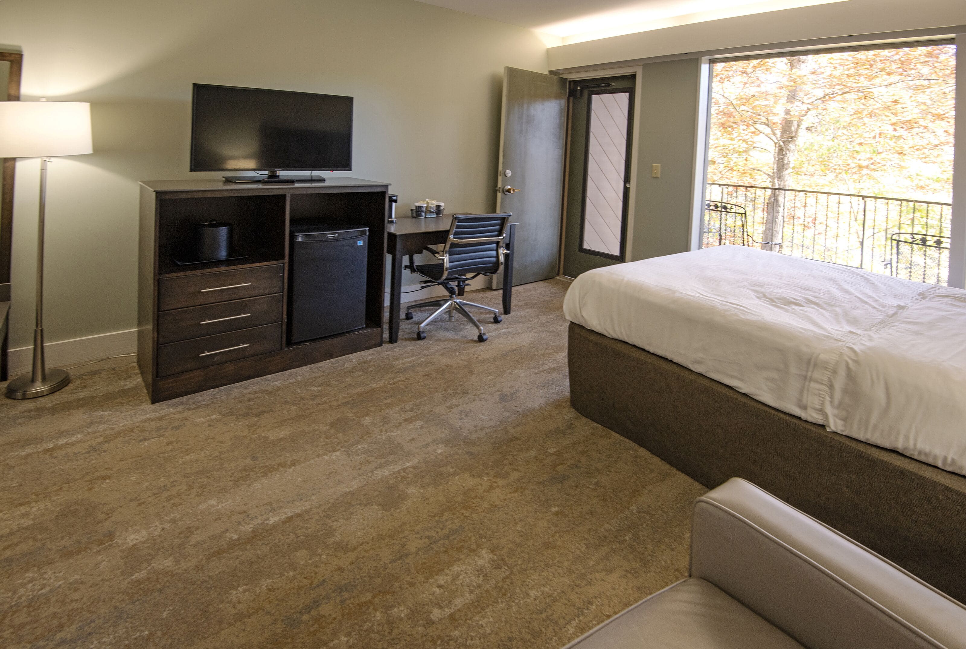 A hotel room with a bed, a desk with a chair, a television, and a door leading to a balcony with a view of trees.