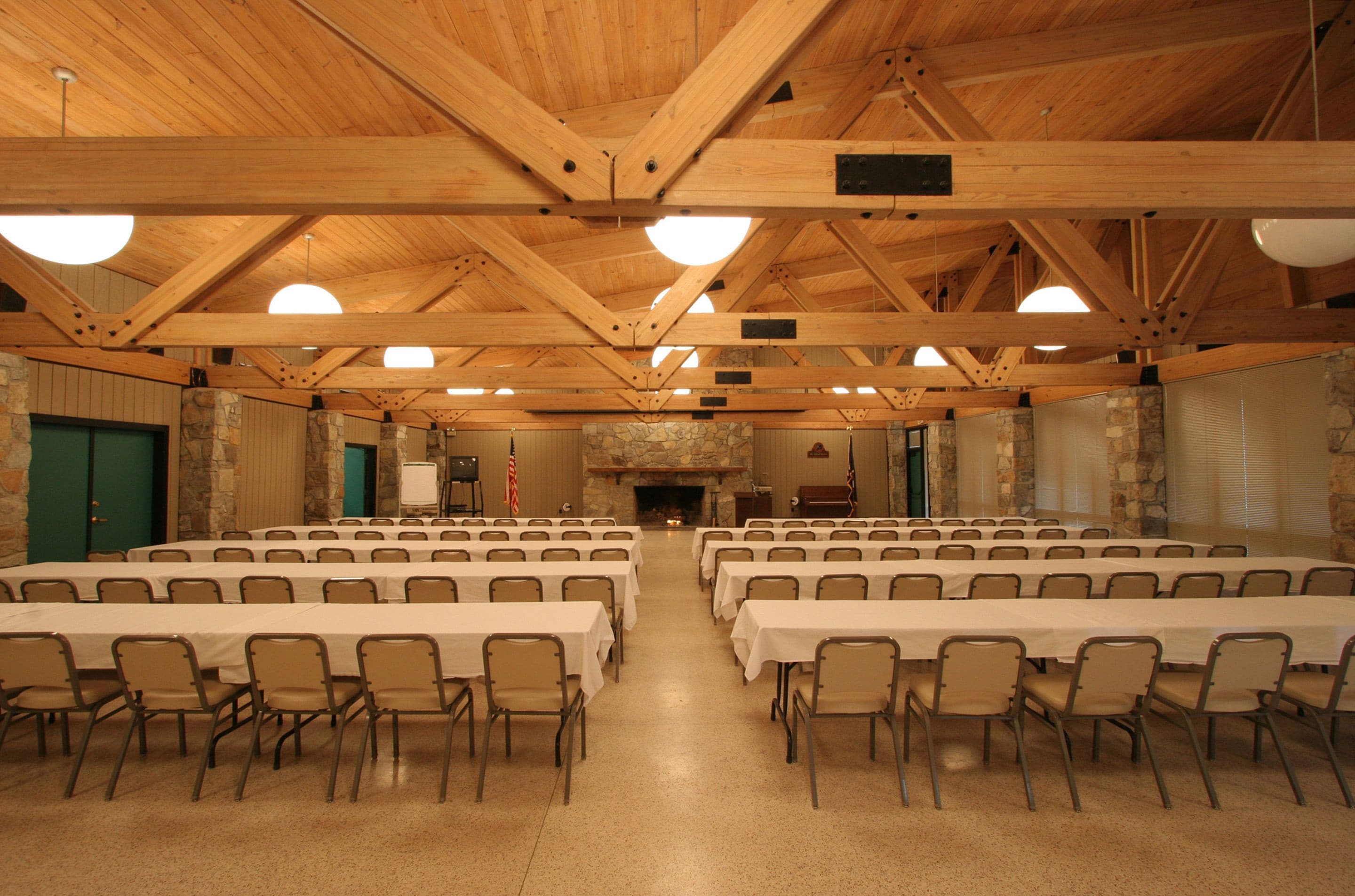 A large hall with multiple tables and chairs, wooden beams on the ceiling, and a stone fireplace.