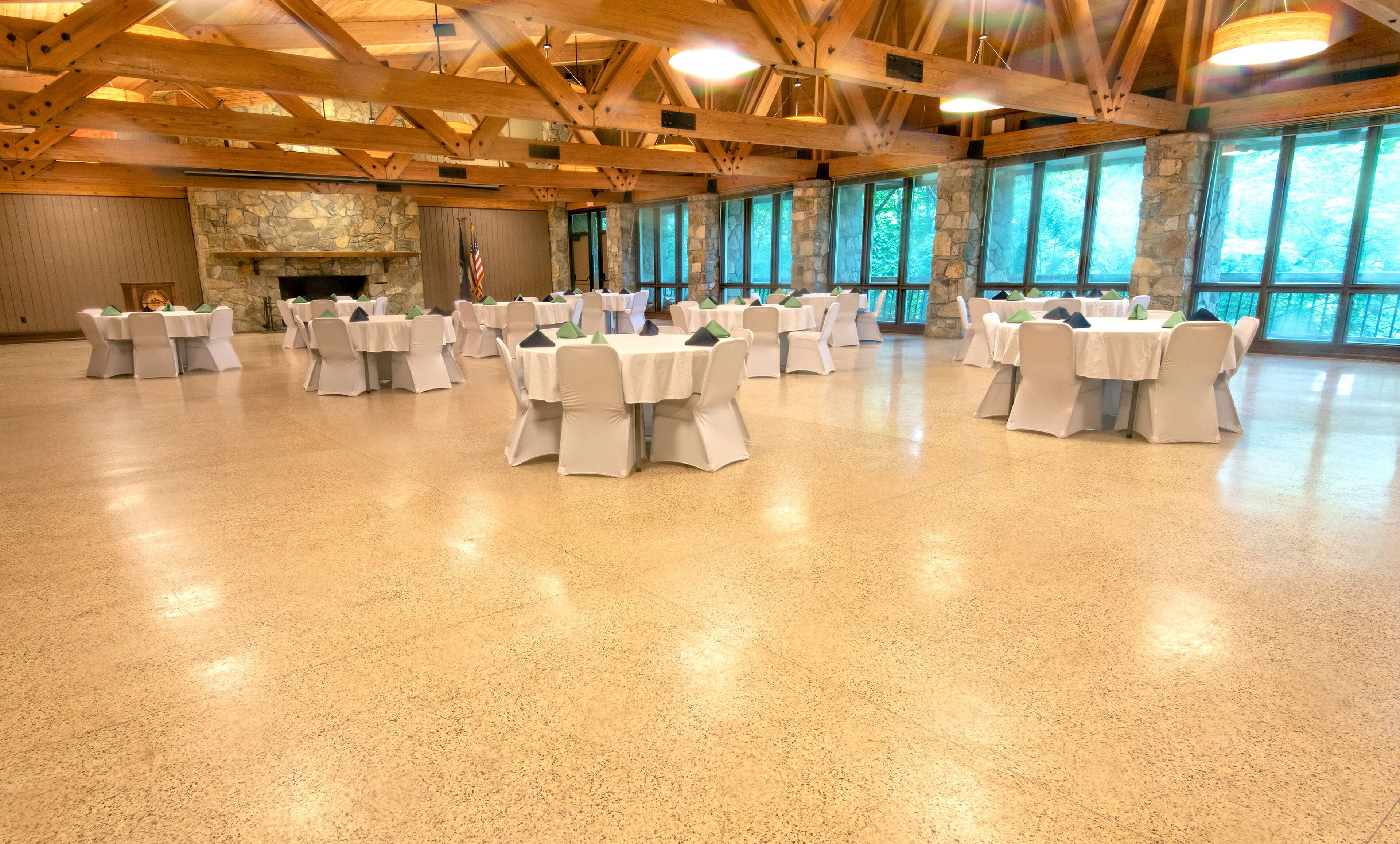 A spacious hall with multiple round tables and chairs, a stone fireplace, and large windows with a view of trees.