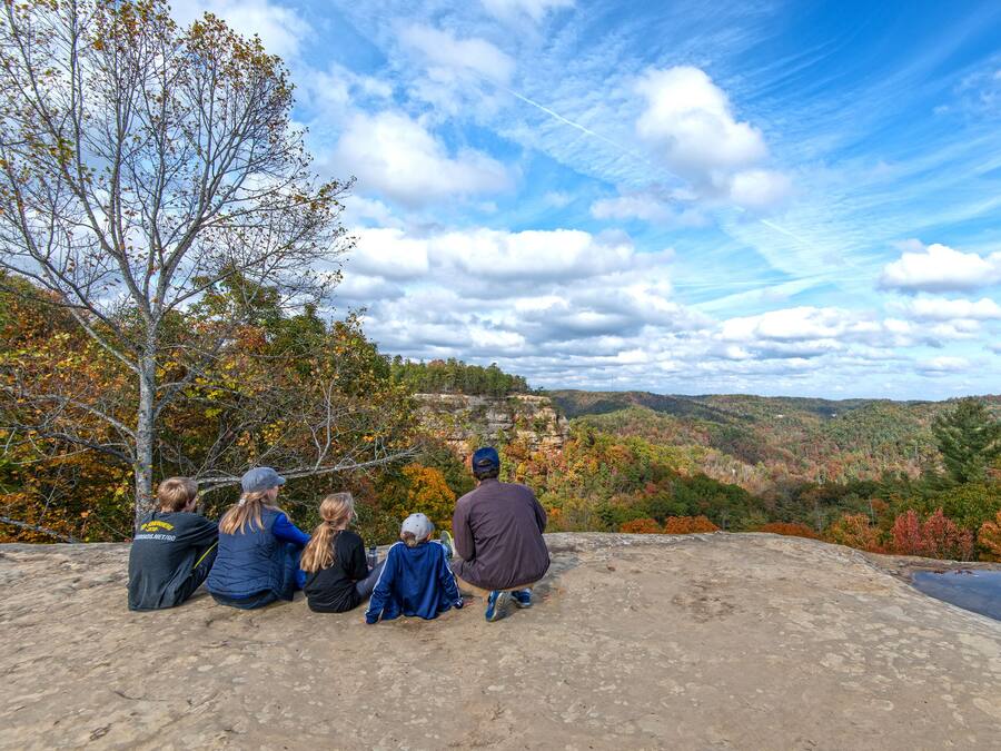 A group of people sitting on a concrete ledge overlooking a scenic landscape with autumn foliage and a clear sky.