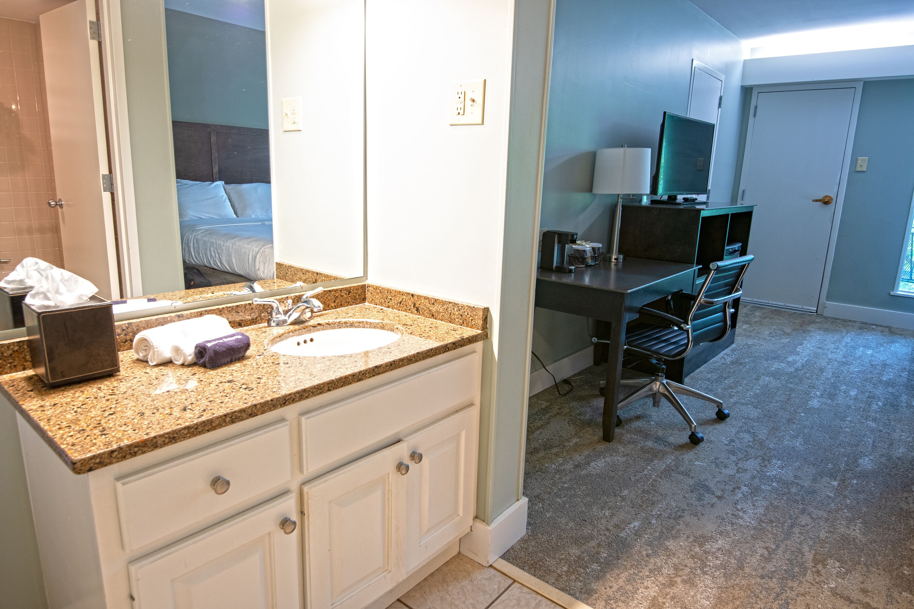 Habitación de hotel con baño que cuenta con lavamanos de encimera de granito, un espejo y tocador blanco. Hay un escritorio con computadora y una silla, y una puerta que da a otra habitación.
