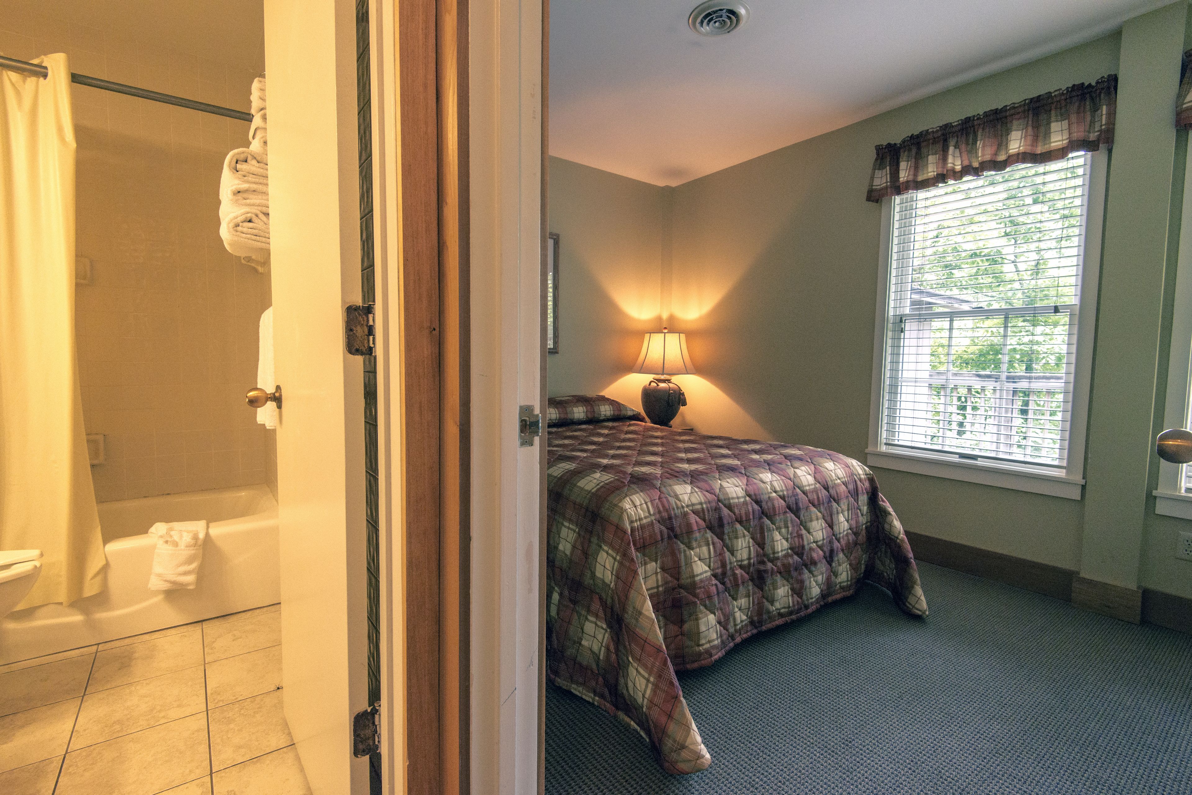 Un dormitorio con cama, ventana con persianas, lámpara y baño con cortina de ducha y toalla colgada en la pared.