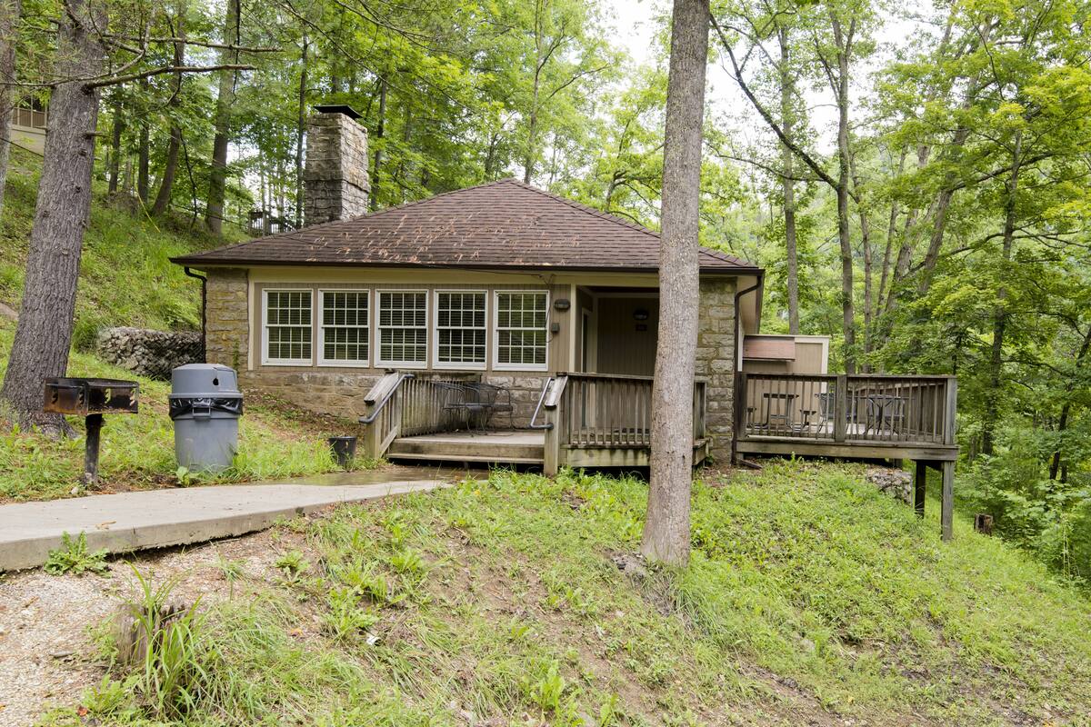 A rustic cabin with a stone chimney, a wooden deck, and a concrete pathway leading to the entrance.
