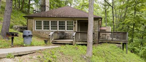 A rustic cabin with a stone chimney, a wooden deck, and a concrete pathway leading to the entrance.