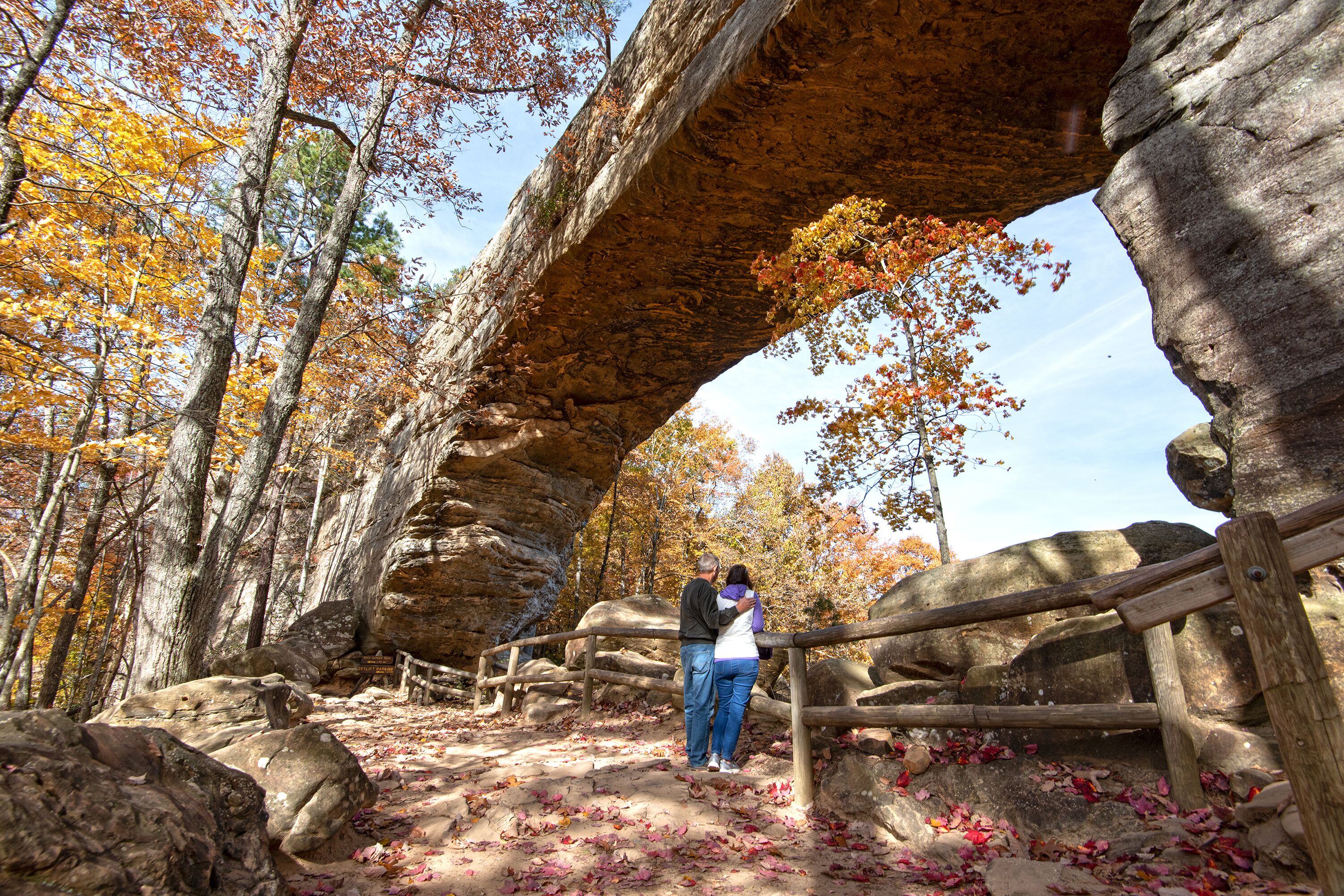 A couple standing under a rock bridge in a forested area with autumn leaves on the ground.