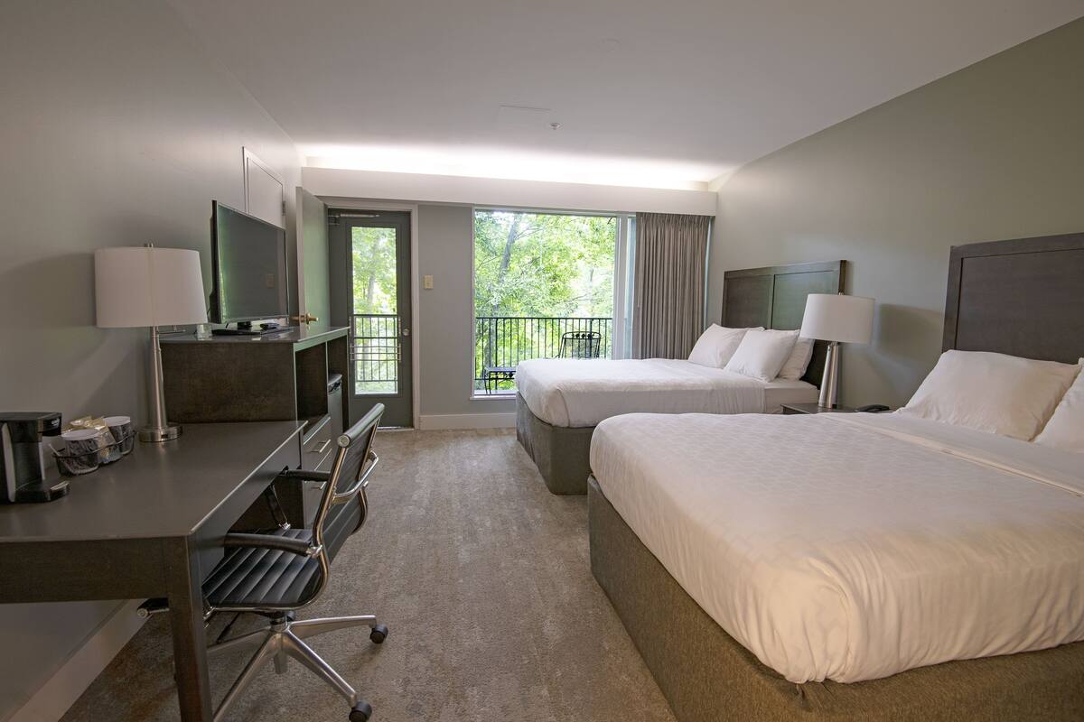 A hotel room with two beds, a desk with a chair, a TV, and a balcony with a view of trees.