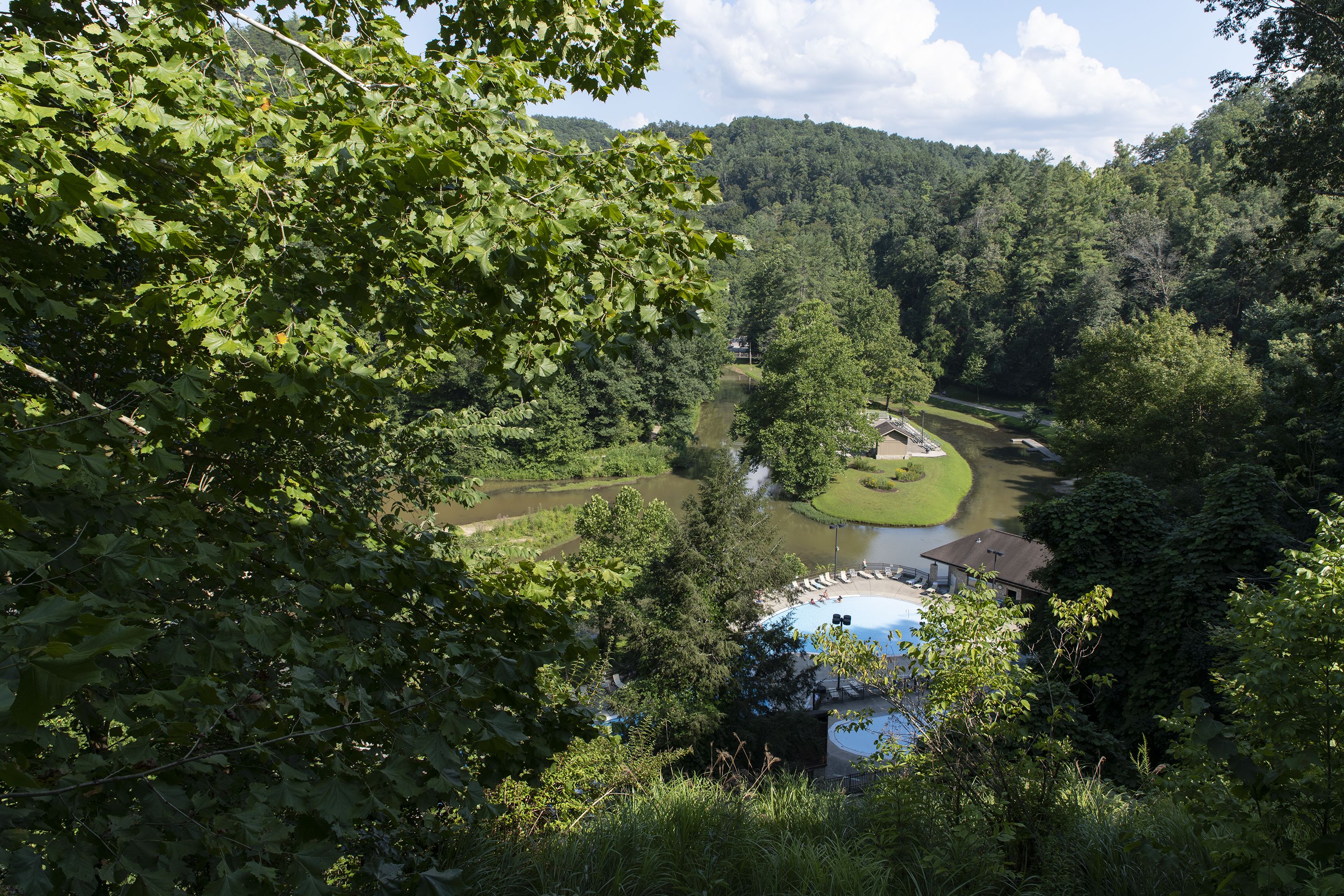 A view of a lush green valley with a river, surrounded by dense forest. There is a swimming pool area with a building nearby.