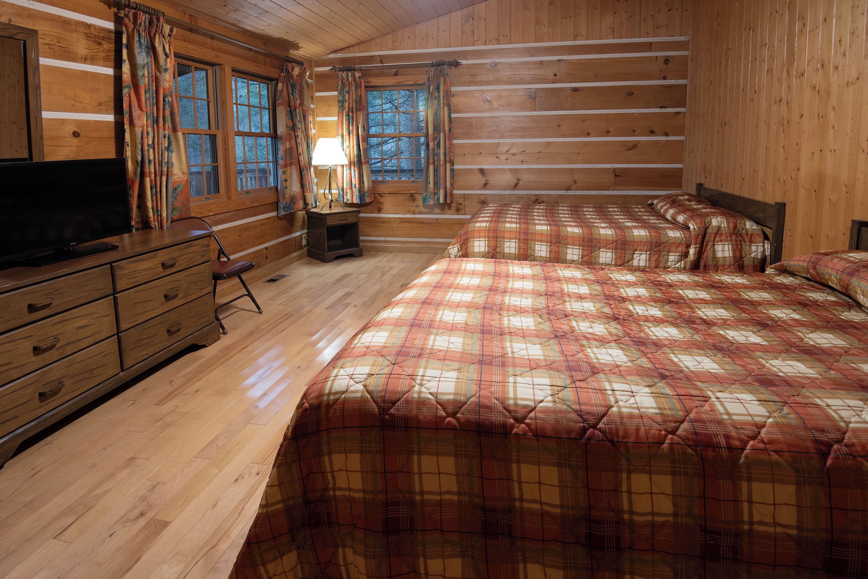 A log cabin bedroom with two beds, a TV, wooden furniture, and large windows.