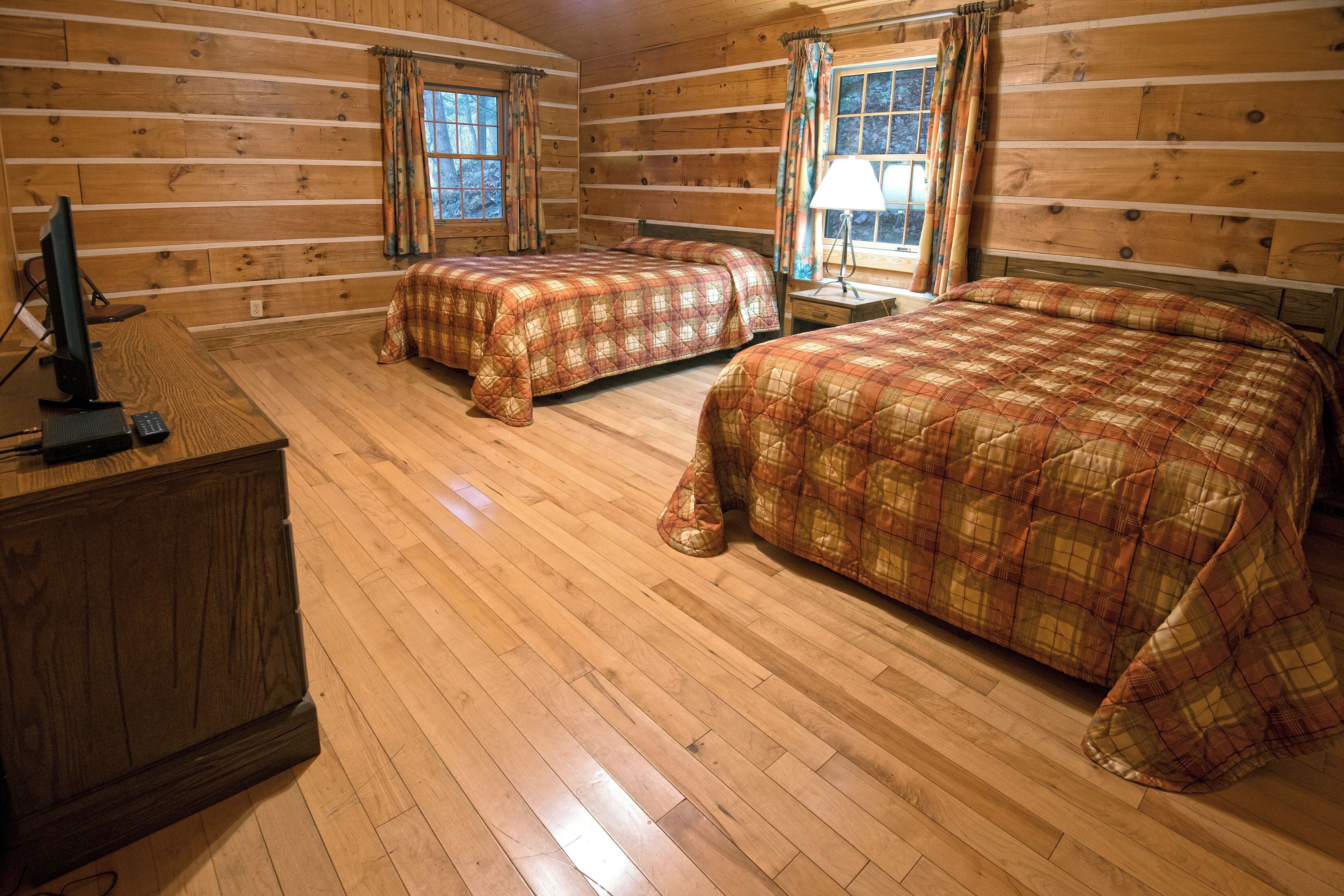 A log cabin bedroom with two beds, a desk with a computer, and a window with curtains.
