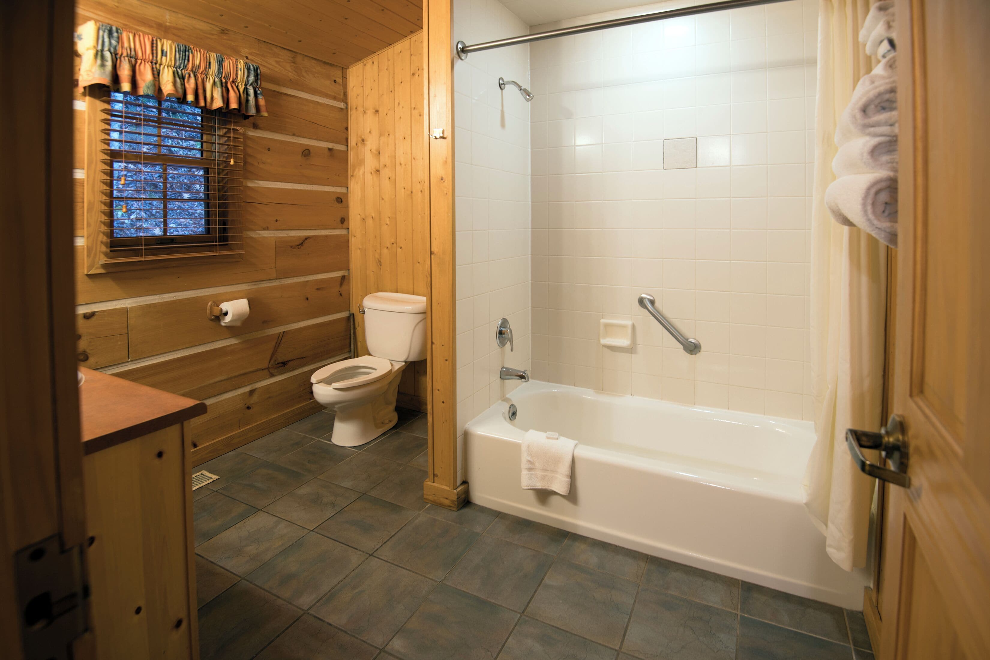 Een houten badkamer met een badcabine, toilet en raam met gordijn.