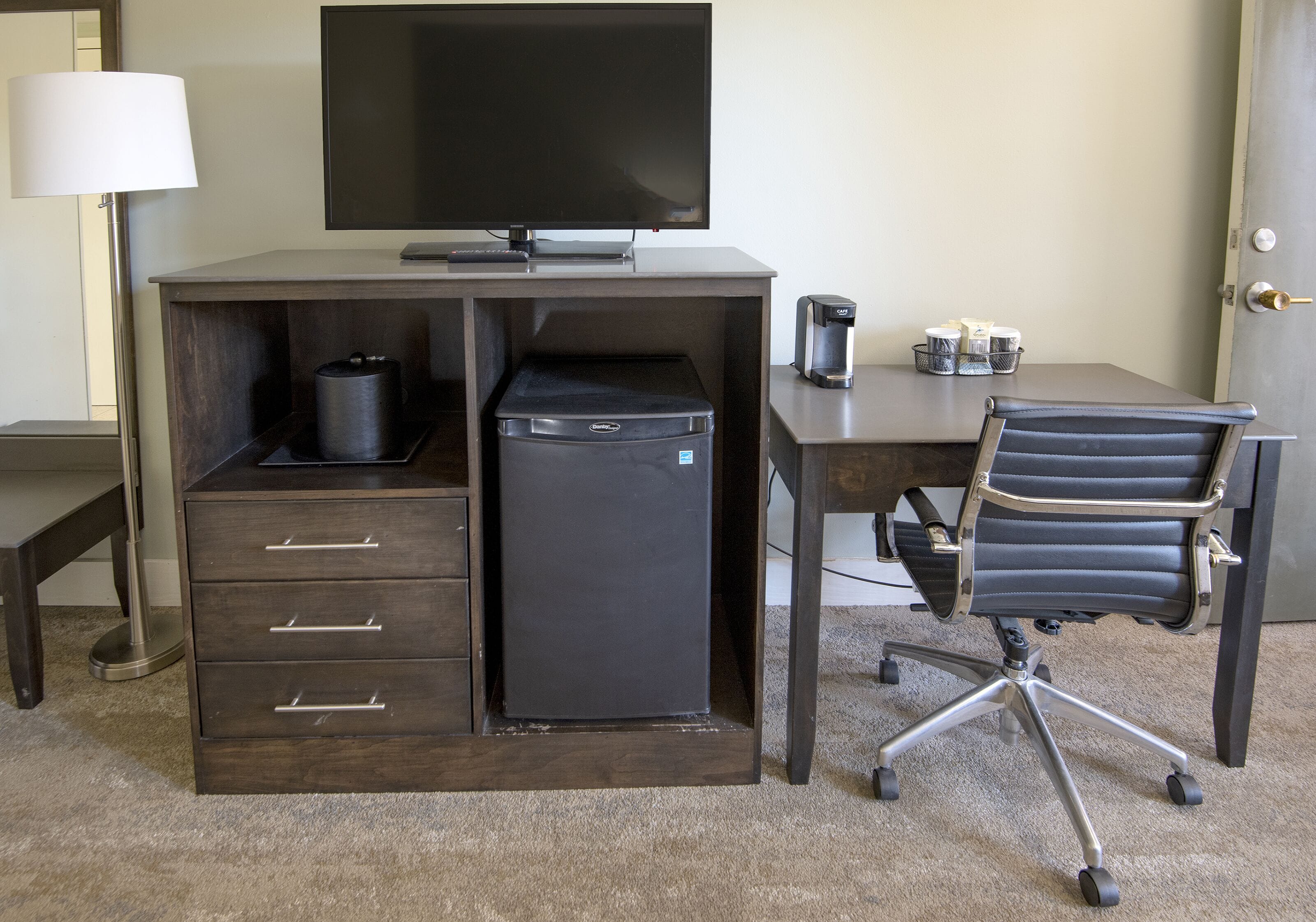 A compact hotel room with a dark wood desk, a black office chair, a small refrigerator, a coffee maker, and a flat-screen TV.