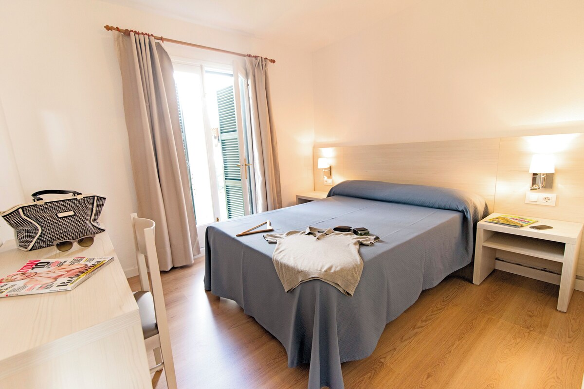 Una habitación de hotel con cama, mesitas de noche, escritorio y silla, y ventana con cortinas.