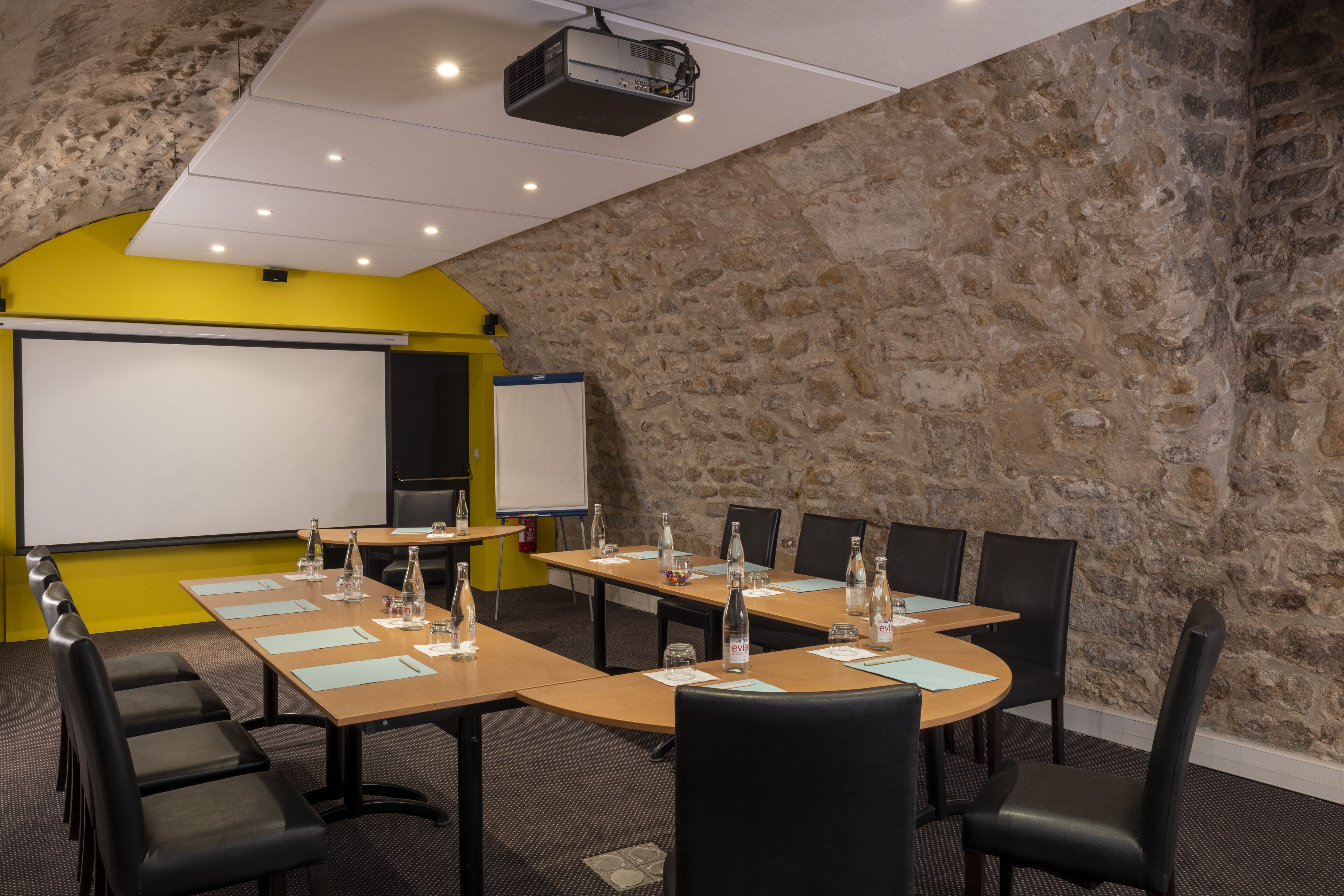 Un salón de conferencias con pared de piedra de fondo, una pizarra y un proyector. Las mesas están dispuestas con botellas de agua y blocs de notas.