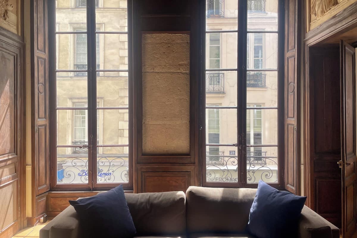 Une pièce dotée d’une grande baie vitrée, d’un plancher en bois et d’un canapé beige agrémenté de deux coussins bleus.