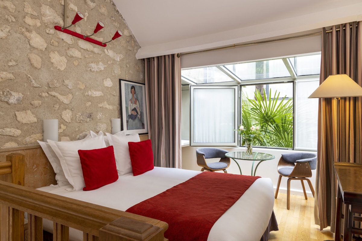 Habitación de hotel con una cama grande, dos sillas, una mesita, una lámpara y vistas a zonas verdes a través de la ventana.