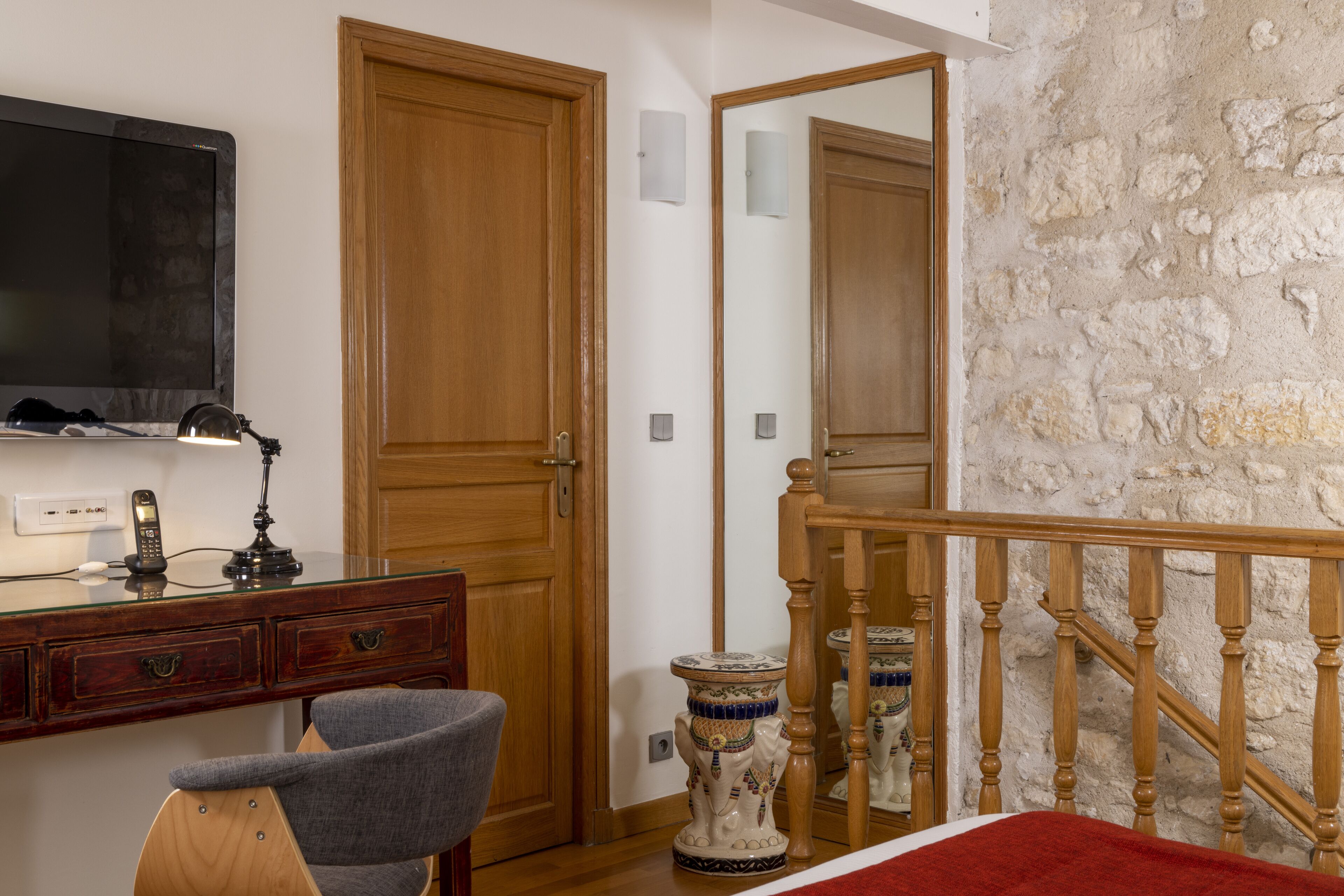 A room with a wooden desk, a chair, a bed with a red blanket, a large mirror, and a stone wall.
