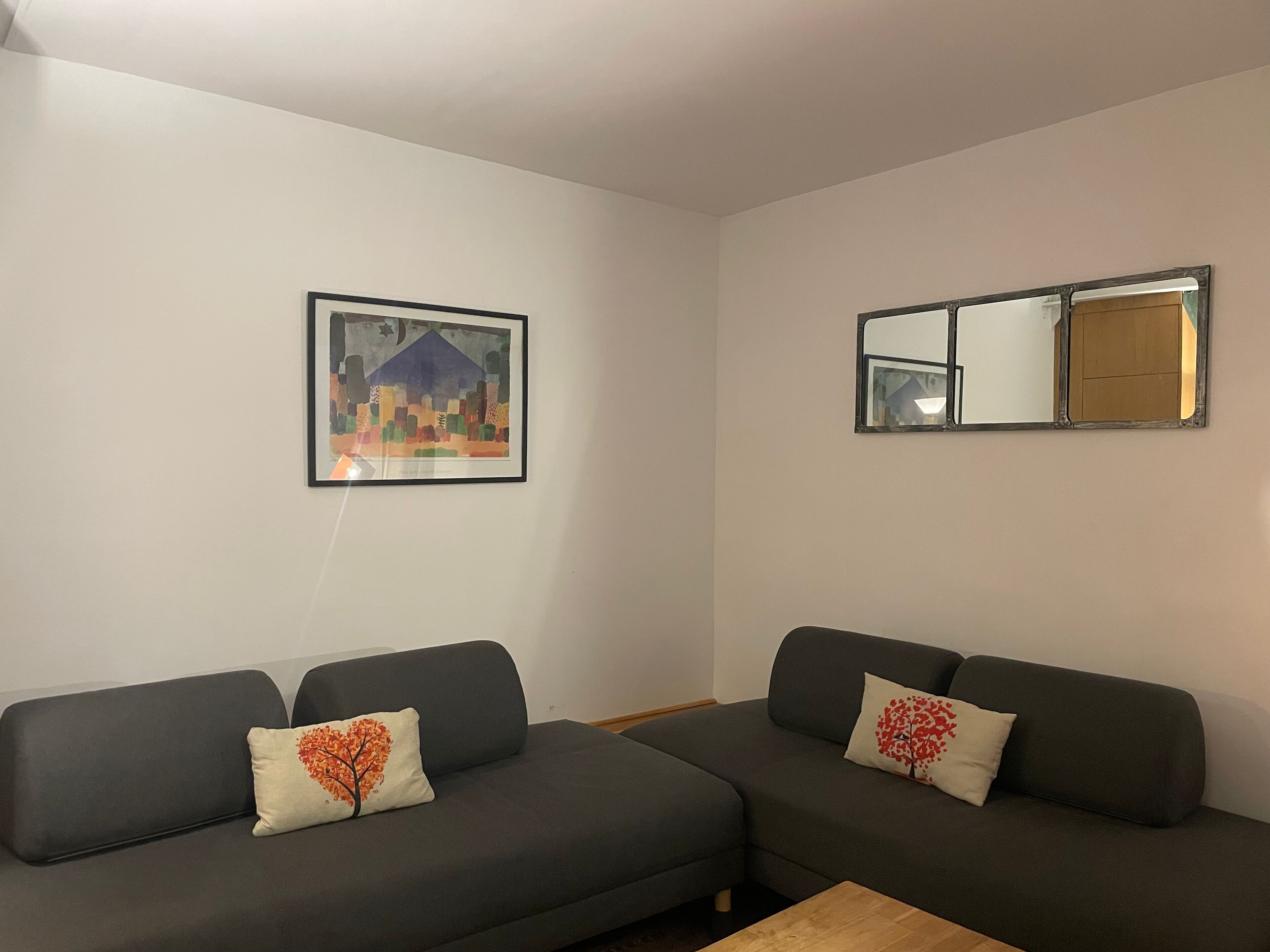 Un salon moderne avec deux canapés gris foncé, chacun agrémenté d’un coussin à motifs d’arbres rouges. Un tableau représentant un paysage urbain, encadré, est accroché au mur, et un miroir est fixé au-dessus des canapés.