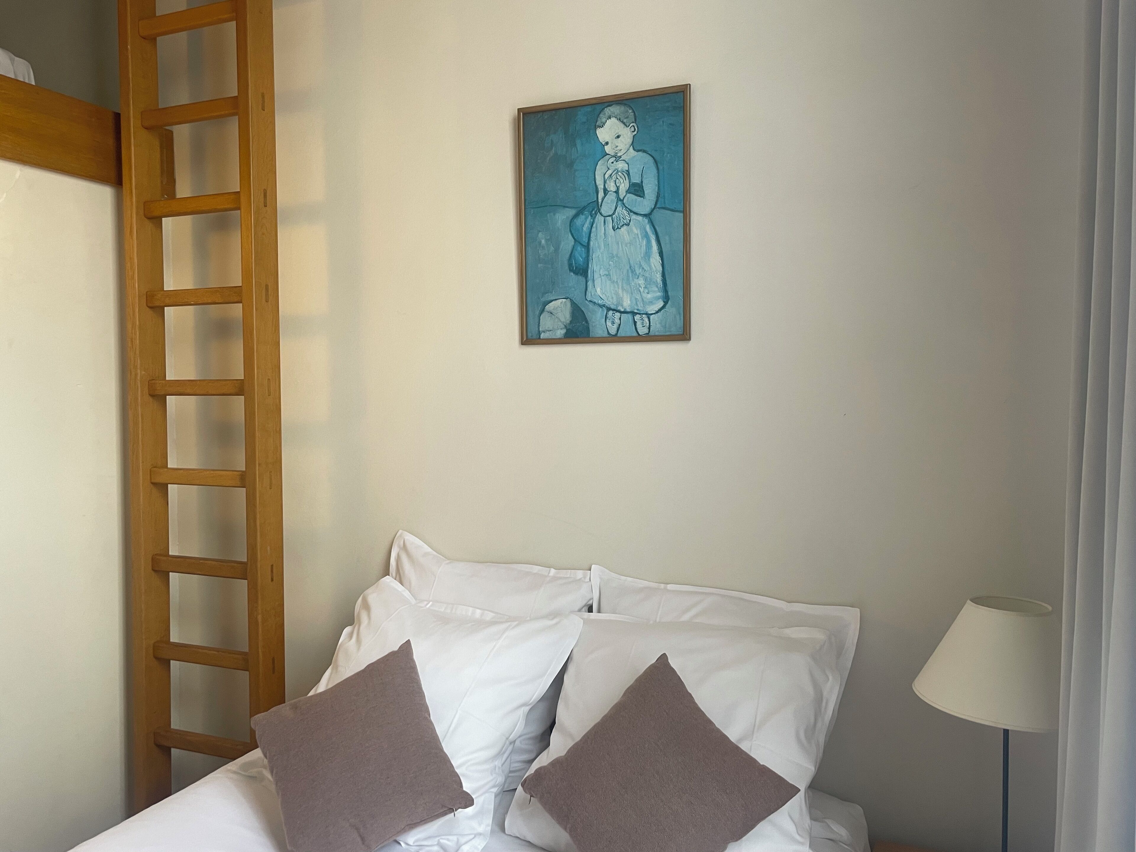 Un dormitorio con una escalera de madera, una cama con almohadas blancas y marrones, un cuadro en la pared y una lámpara en la mesita de noche.