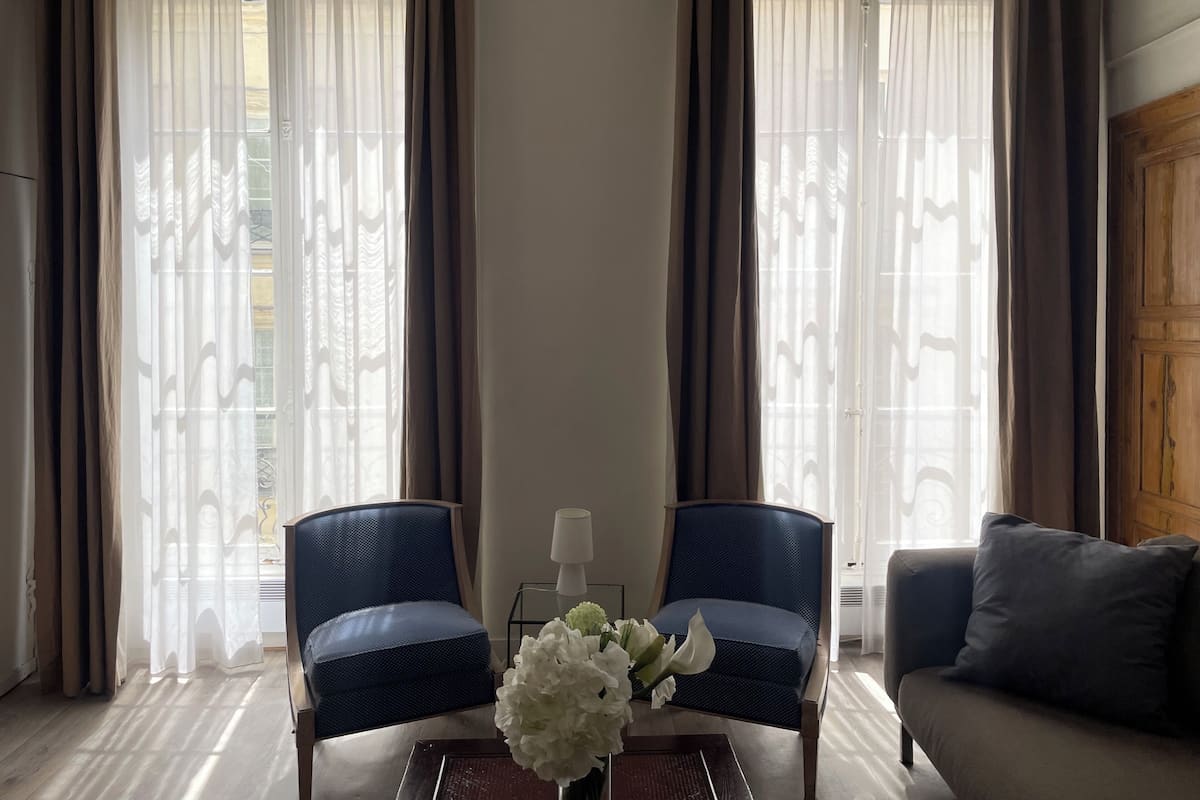 Une pièce avec deux chaises bleues, une petite table sur laquelle est posé un vase de fleurs blanches, et un canapé gris.