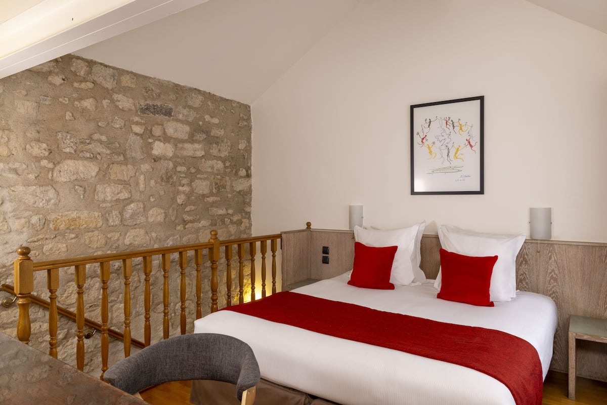 Une chambre avec un mur en pierre, un escalier en bois, un lit avec une literie blanche et rouge, et une œuvre d’art encadrée accrochée au mur.