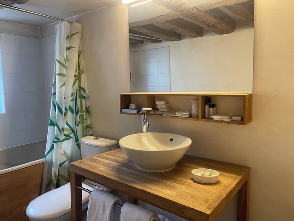 Un baño con tocador de madera, un lavamanos grande, un inodoro y una ducha con cortina de bambú.