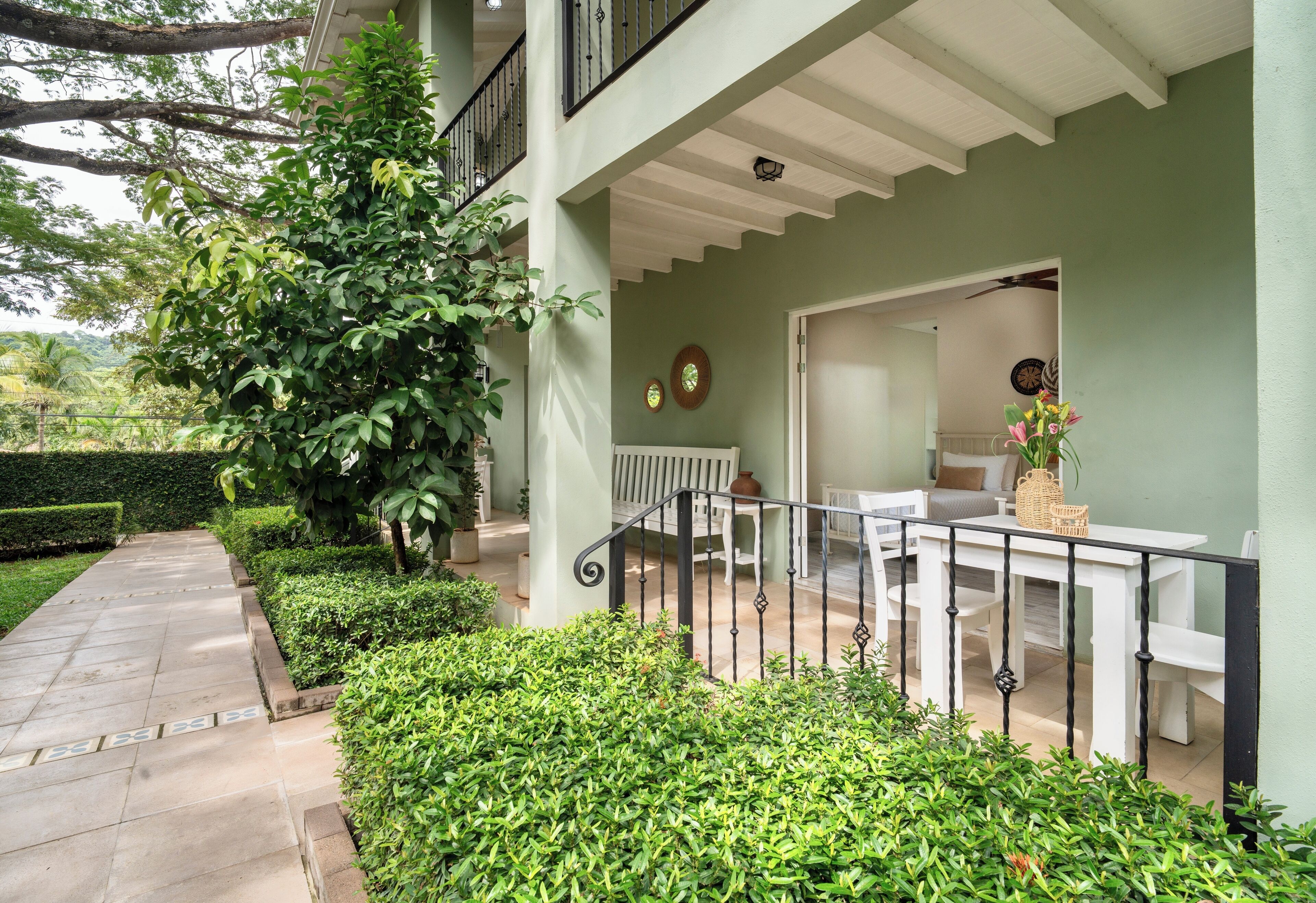 A covered outdoor area with a white bench and table, surrounded by greenery and a stone pathway.