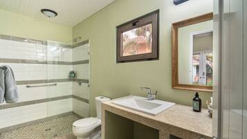 A bathroom with a glass shower enclosure, a toilet, a sink with a mirror, and a window with a view of palm trees.
