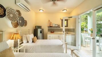 Habitación con dos camas, ventilador de techo, aire acondicionado, una mesa con lámpara, refrigerador, área de cocina con pileta y alacenas, y puerta corrediza de vidrio que da a un espacio exterior.