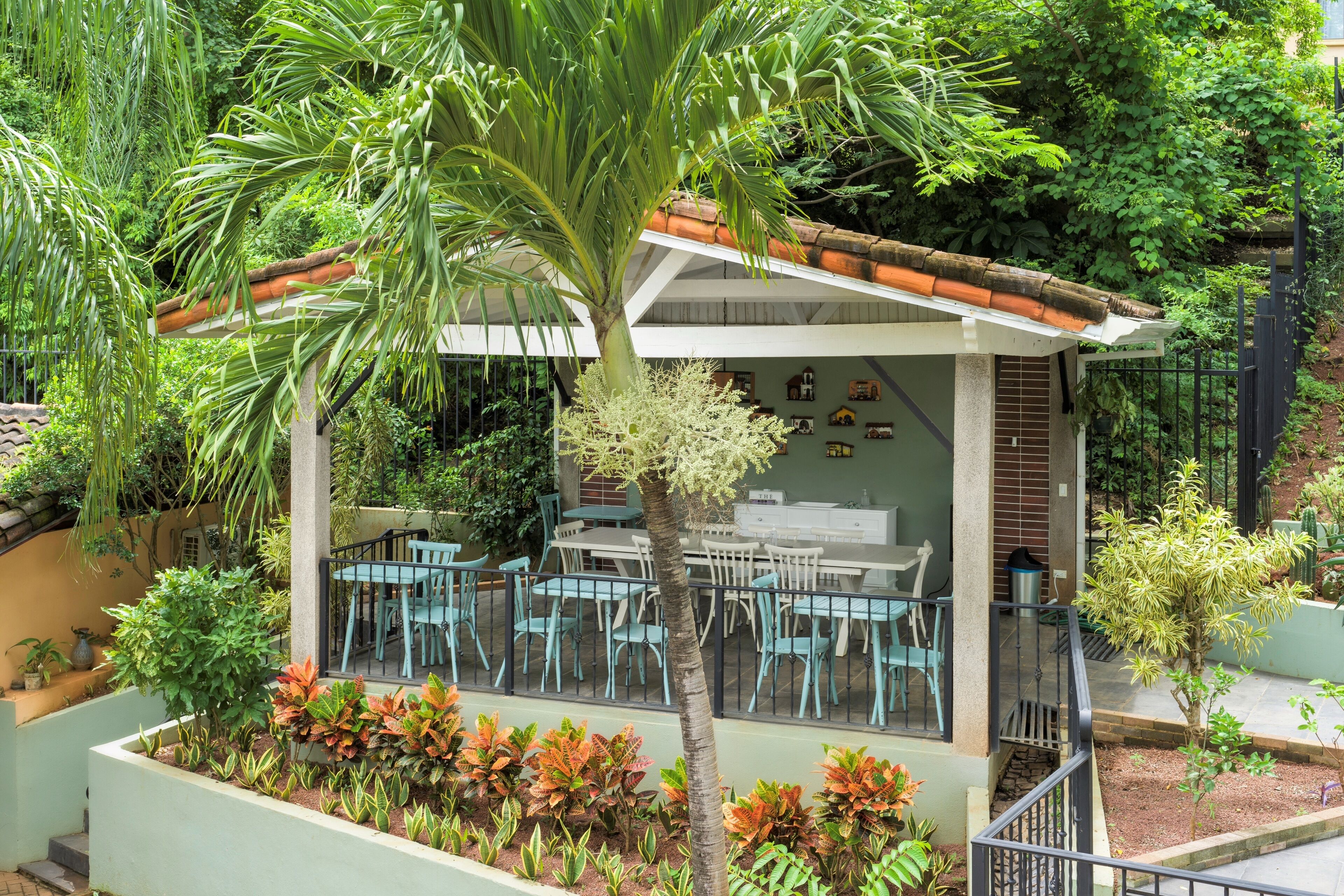 A covered outdoor seating area with tables and chairs, surrounded by various plants and a palm tree.