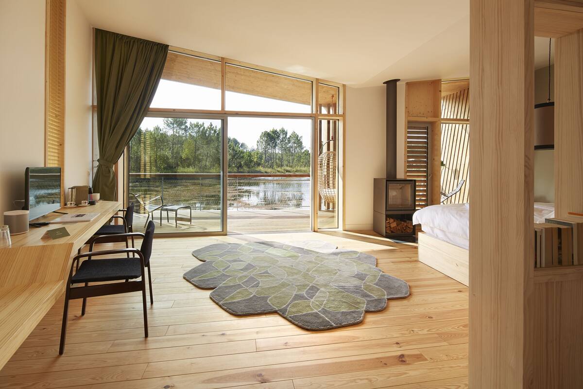 A modern bedroom with a large window, wooden floor, a bed, a desk with a chair, and a view of a lake.
