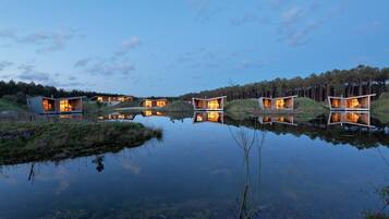 A series of modern, wooden cabins with large windows, situated on a lakeside with reflections.