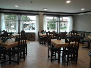 Uma sala de jantar com várias mesas e cadeiras, janelas amplas com cortinas e vista para a rua.