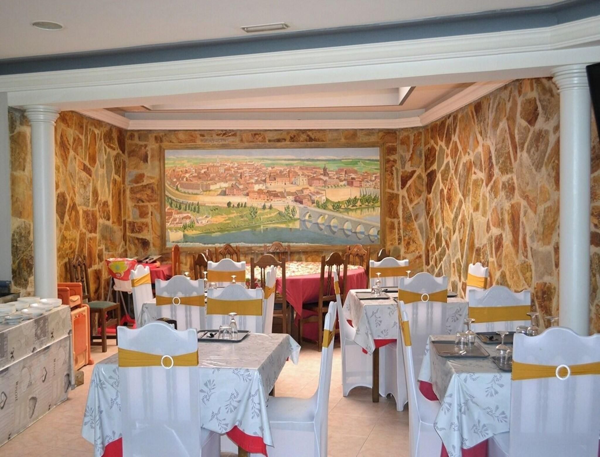 Un restaurante con paredes de piedra, un cuadro grande de un pueblo y mesas con sillas blancas y cintas amarillas.